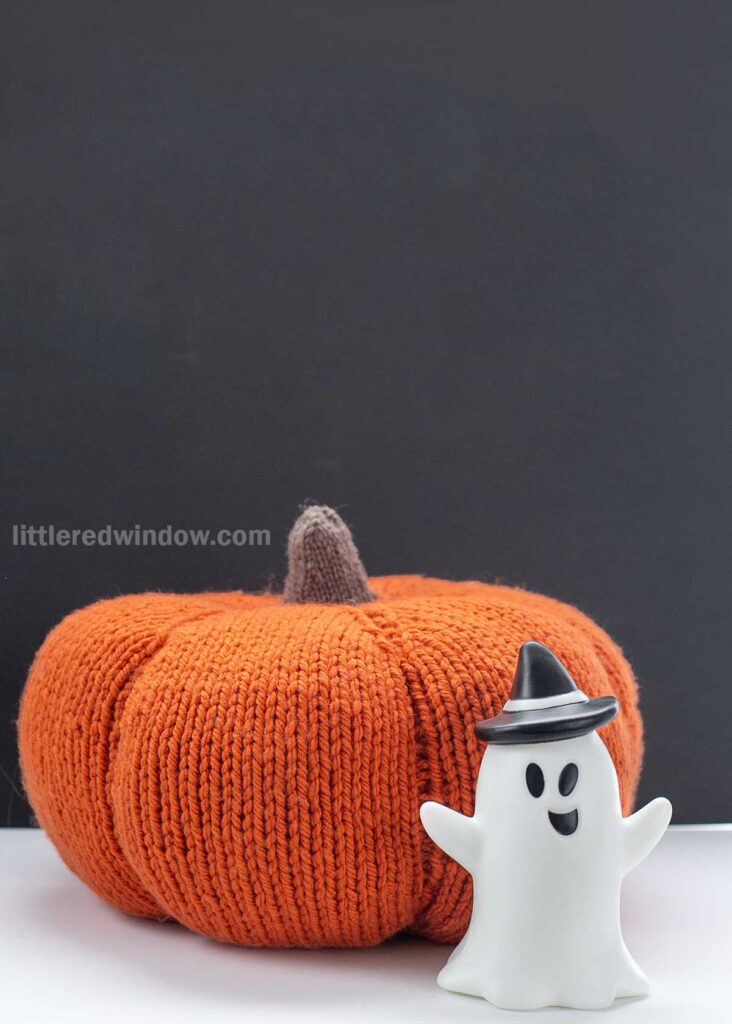 A knitted orange pumpkin and a small plastic ghost figurine with a black hat are placed against a plain dark backdrop. The pumpkin has a brown knitted stem, and the ghost is smiling with its arms raised. The text