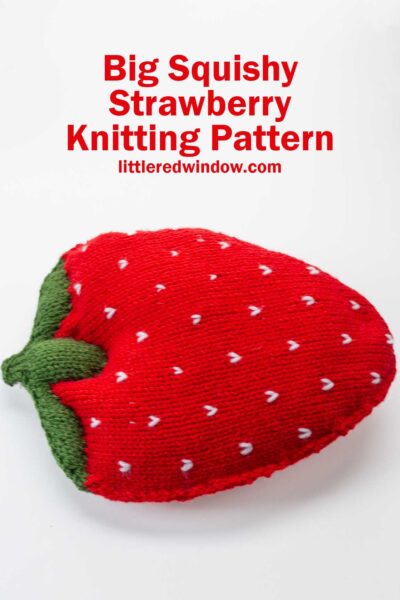 A large, plush knitted pillow shaped like a strawberry with green leaves and white seed details. Red text above reads: "Big Squishy Strawberry Knitting Pattern littleredwindow.com" on a white background—perfect for strawberry knitting pattern lovers.