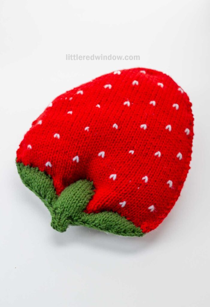 A knitted red pillow designed to look like a strawberry, featuring a charming strawberry knitting pattern with green leaves at one end and small white stitched dots representing seeds.