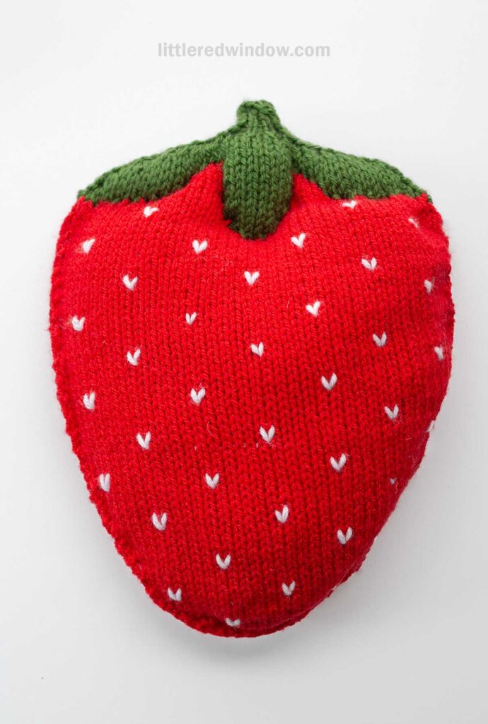 A red, knitted object shaped like a strawberry with green at the top and white embroidered seeds, crafted using a strawberry knitting pattern and displayed on a white background.