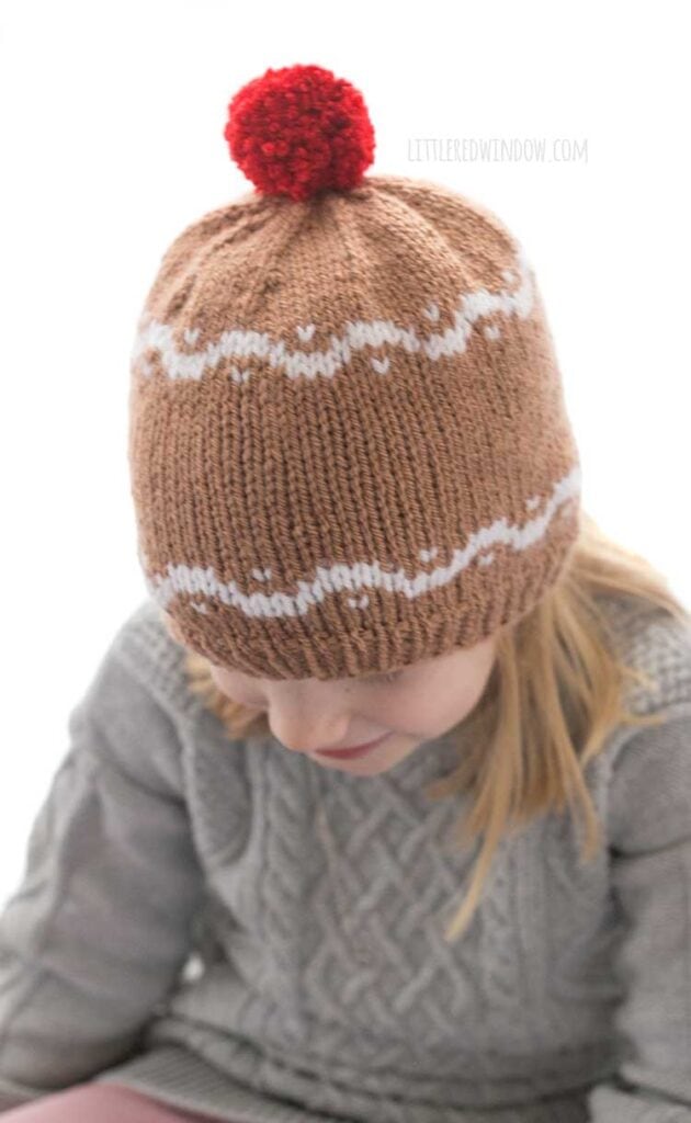 girl in gray sweater wearing a medium brown hat with white designs that look like gingerbread and a red pom pom on top looking down
