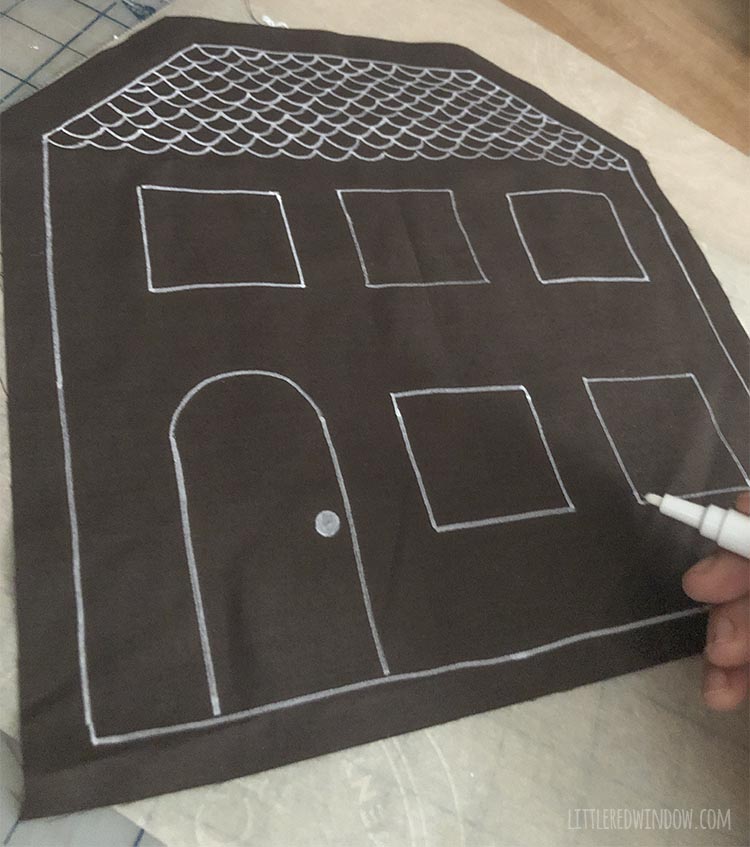 a hand holding a white pen drawing frosting decorations on a brown fabric gingerbread house shape