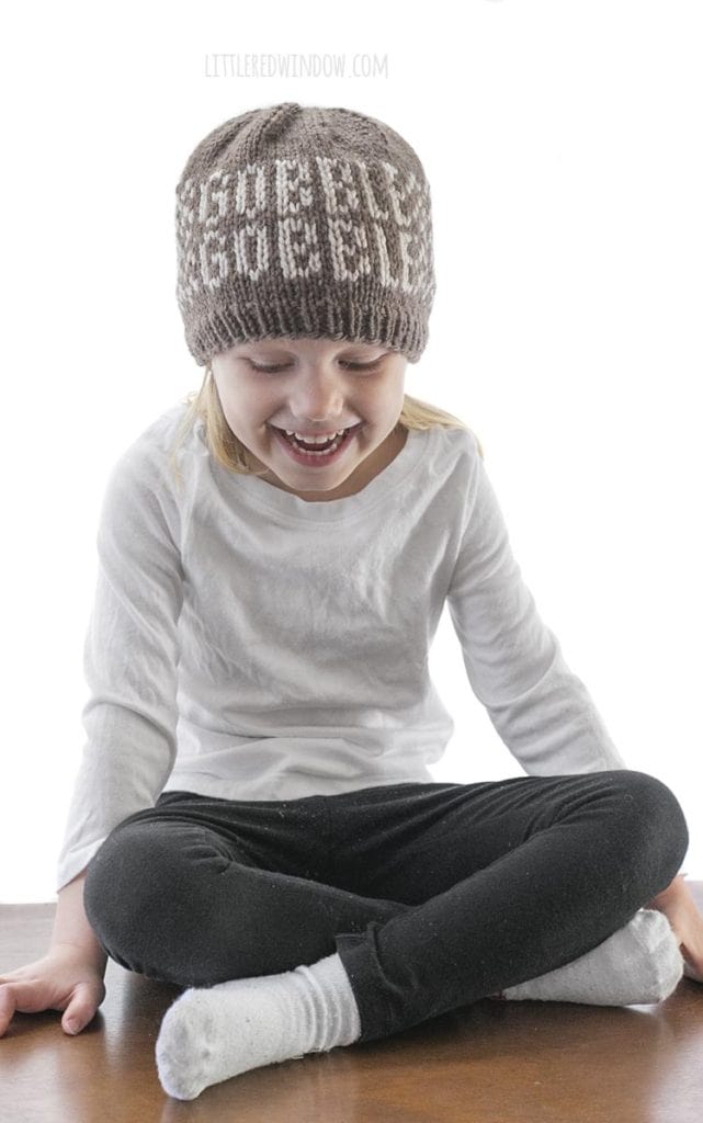 little girl sitting cross legged and wearing black pants white shirt and brown knit hat that says gobble gobble on the front