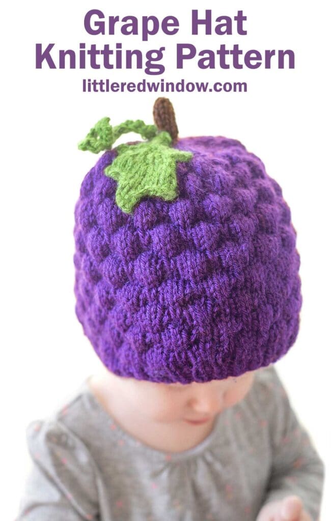 A baby is wearing a purple knitted grape hat, complete with a green leaf and stem on top. The text above reads, 