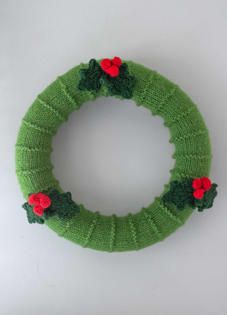 A knitted green Christmas wreath is hanging on a wall, showcasing a delightful holly wreath knitting pattern. It features three sets of red berries and green holly leaves as decorations against a plain light gray background.