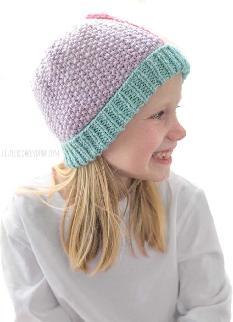 Smiling girl in white shirt looking off to the far right and pink pants wearing a knit hat that has a light teal ribbed brim and is half light purple seed stitch on the left