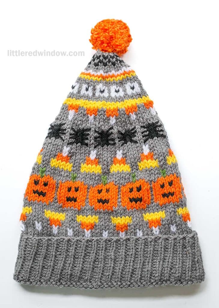 A Halloween stocking cap with themed patterns, featuring pumpkins, ghosts, spiders and candy corns in orange, black, white, and yellow colors. The hat is primarily gray with a bright orange pompom on top.