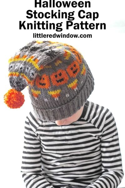 A child wearing a black and white striped shirt looks down, showcasing a gray knit Halloween stocking cap adorned with orange and black pumpkin patterns, candy corns, spiders and an orange pom-pom. The text above reads, "Halloween Stocking Cap Knitting Pattern" with "littleredwindow.com" underneath.