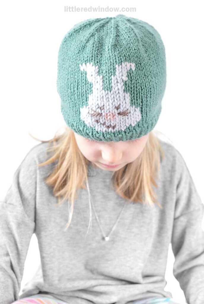 A child wearing a gray shirt and an Easter bunny hat with a charming white bunny design looks downward.