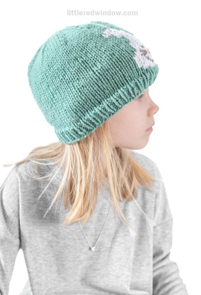 A child with long blonde hair wears a grey shirt and an Easter bunny hat, featuring a teal knit design, looking to the side against a plain white background.