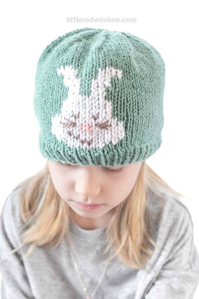 A child wears a green knit Easter bunny hat with a charming white bunny design. With light hair and a gray top, they gaze downward, embracing an adorable moment.