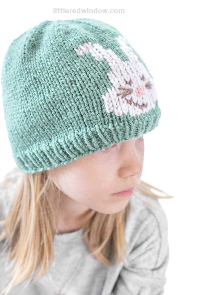 A child wearing an Easter bunny hat—a green knit beanie with a white bunny design—glances slightly to the side. They're dressed in a gray shirt, set against a plain white backdrop.