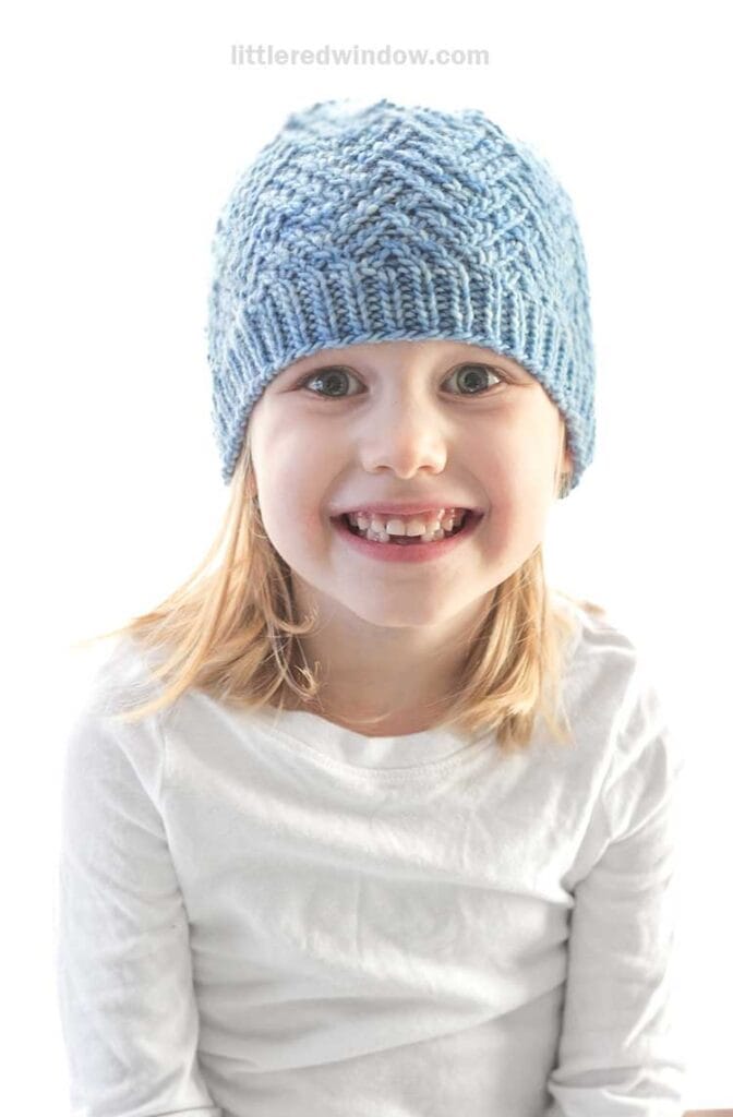 A young child with blonde hair wearing a blue knitted beanie and a white long-sleeve shirt flashes a charming smile at the camera against a crisp white background, making the hashtag-worthy moment unforgettable.