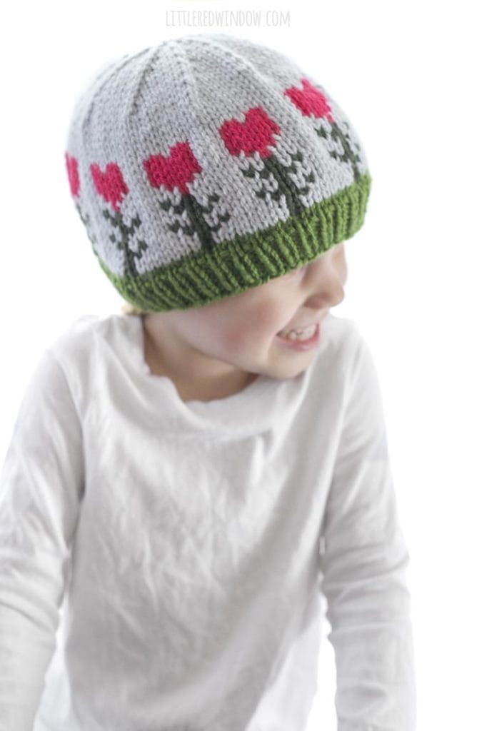 little girl in white shirt wearing light blue knit hat with pink heart shaped flowers on it