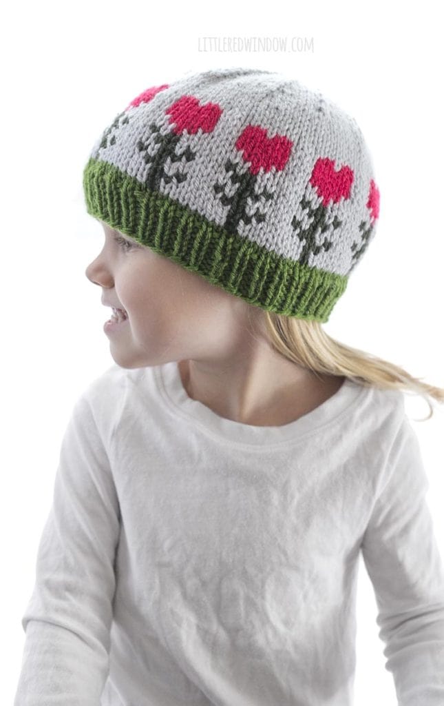 little girl in white shirt wearing light blue knit hat with pink heart shaped flowers on it looking off to the left