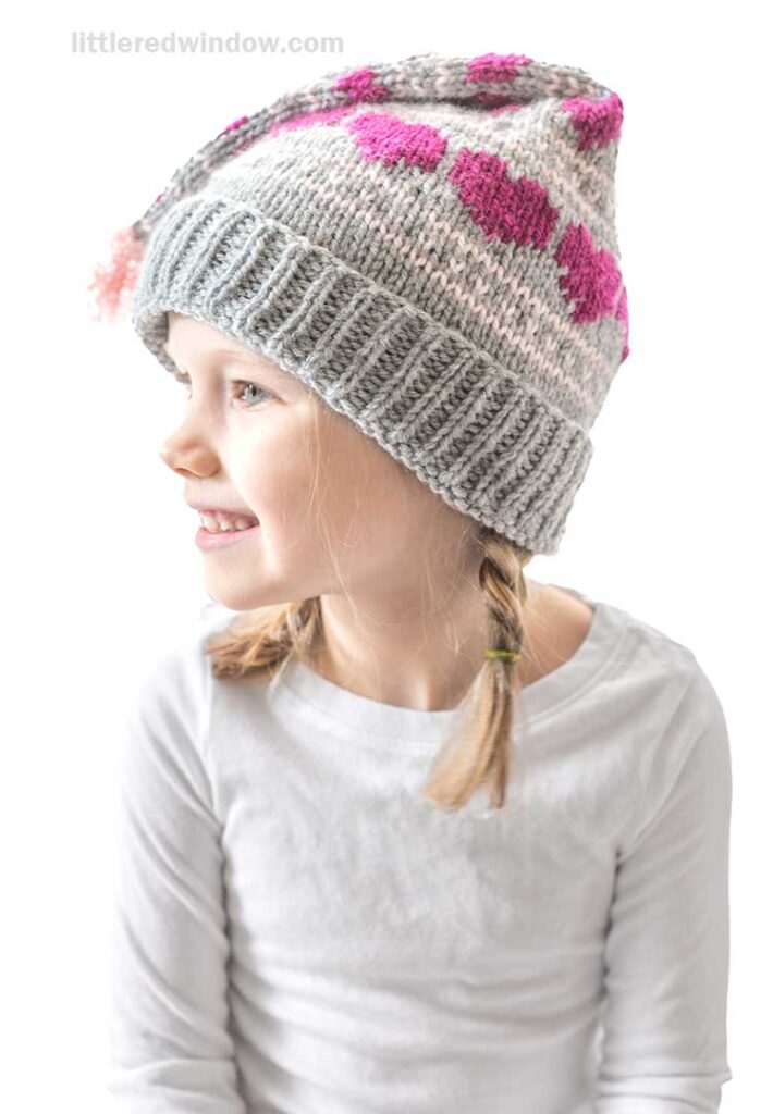 A young girl in a white shirt is smiling and looking to the side. She is wearing a gray knitted hat with pink hearts and a pom-pom, and her hair is styled in two braids.