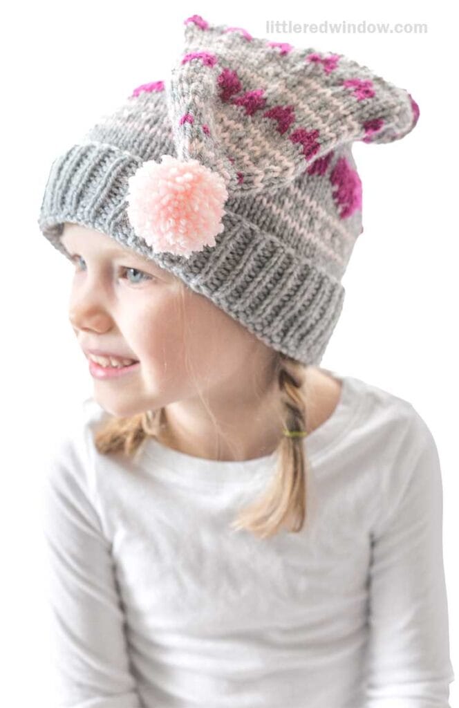 A young girl wearing a gray knitted hat with pink patterns and a pom-pom smiles while facing sideways. She has two braids and wears a white long-sleeve shirt.