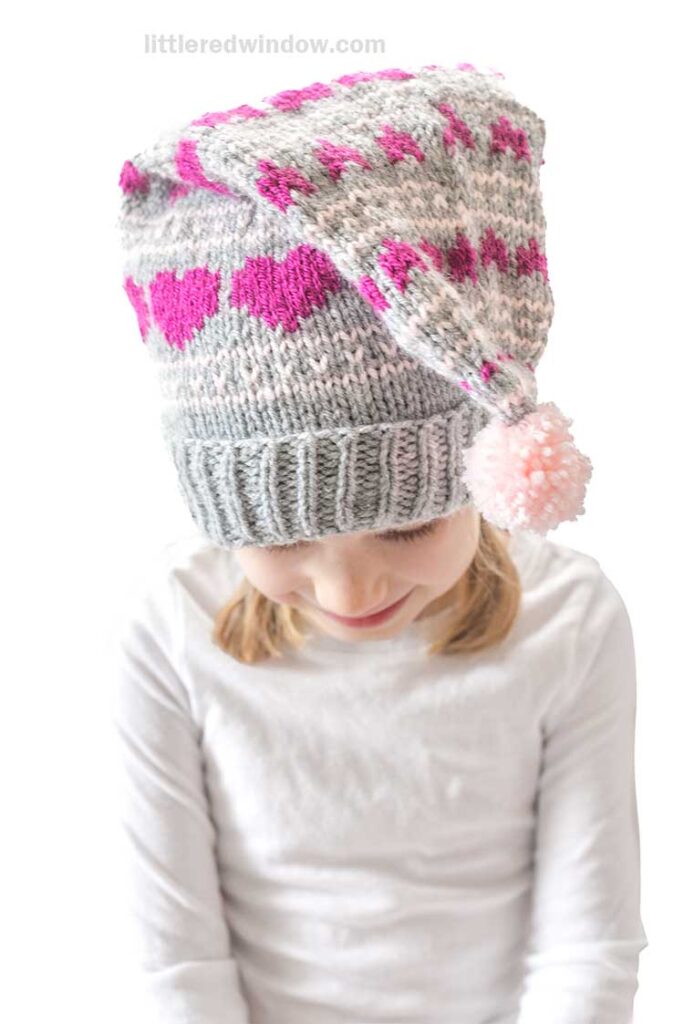 A young girl wearing a white shirt and a knitted hat with pink hearts and patterns. The hat has a pink pom-pom at the tip. The girl is looking down with a slight smile.