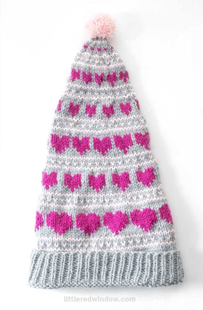 A knitted hat with a long, pointed shape featuring a pattern of pink hearts on a gray background. The hat is topped with a pink pom-pom and has a ribbed edge.