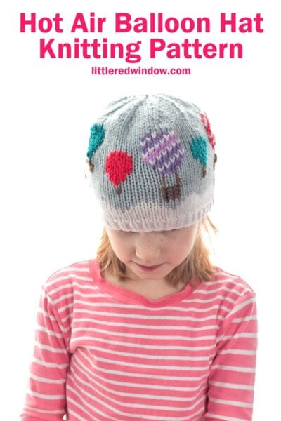 A child in a pink striped shirt wears a knitted hat decorated with colorful hot air balloons. Text above reads,
