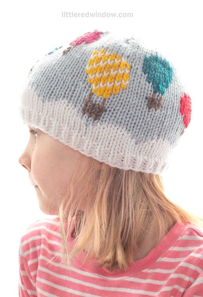 A child wearing a white knit hat with colorful hot air balloon patterns, looking to the side. The child has light hair and is dressed in a pink and white striped shirt.