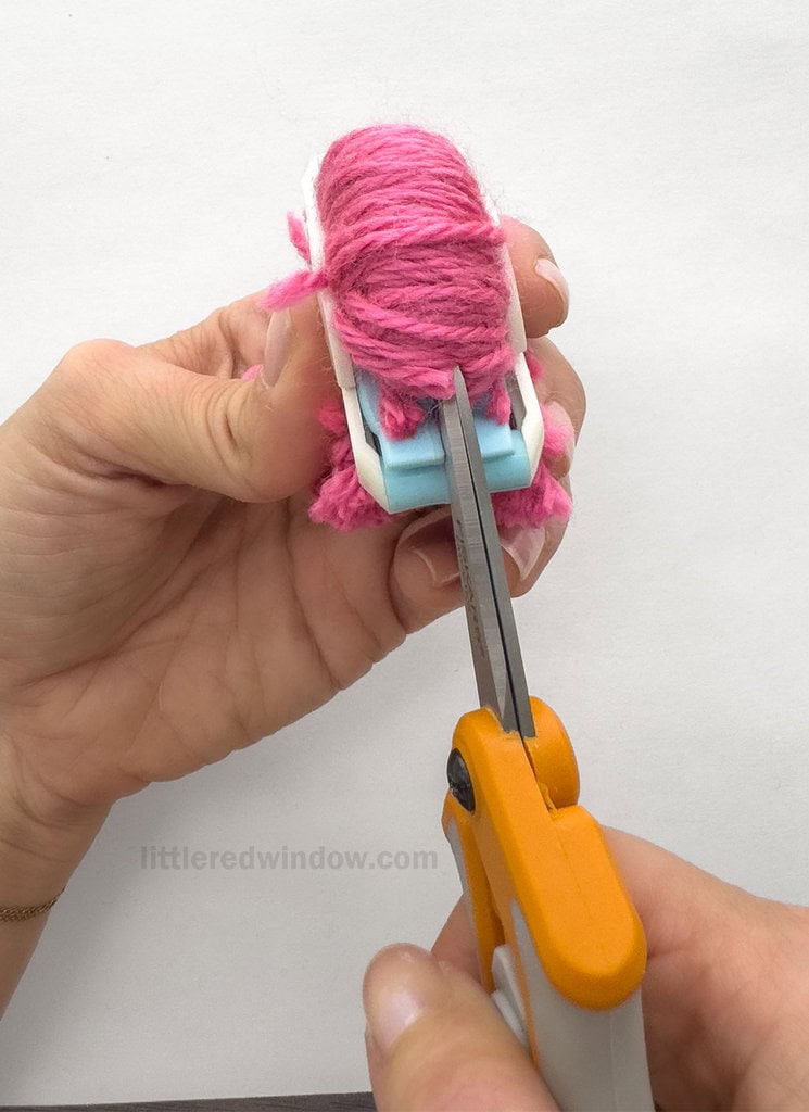 Using the sharpest scissors you can find, and keeping the pom pom maker closed, cut along one edge of one half the pom pom between the gap between the arches, all the way through all of the wrapped layers of yarn.