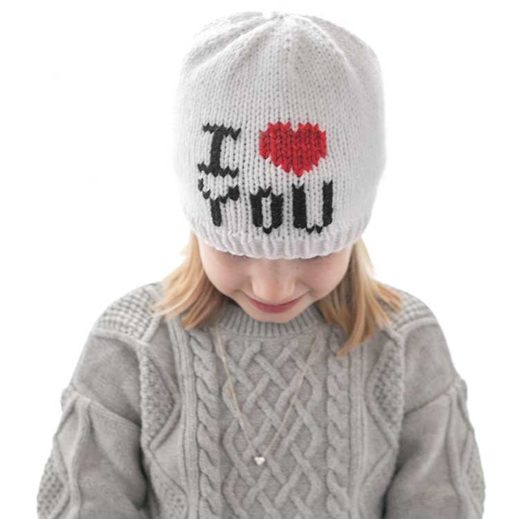 girl in gray sweater dress wearing a white knit hat with the words I heart you where the heart shape is red in front of a white background looking down at her lap
