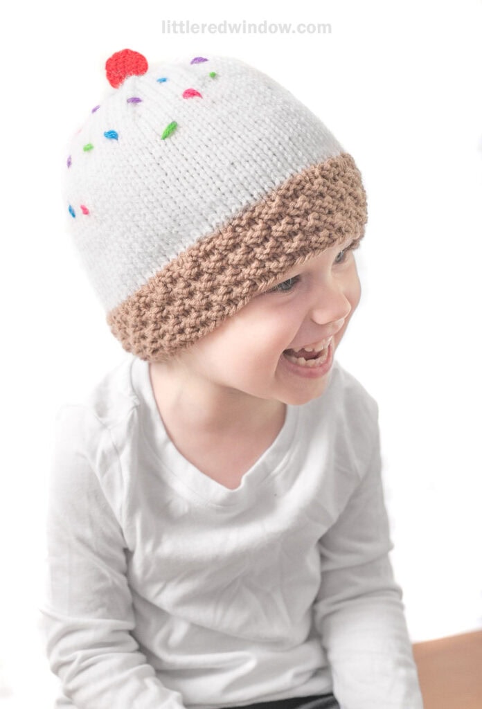A smiling child, wearing a white long-sleeve shirt, is seen facing slightly to the side. The child is donning an adorable ice cream hat designed to look like a cupcake, featuring white frosting with colorful sprinkles and a brown textured brim.