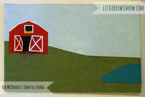 Felt board with a red barn on a green hill, blue sky and small pond