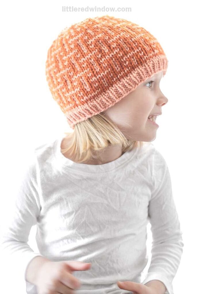 A child in a white shirt wearing an orange knit hat interwoven with a zigzag pattern looks to the side and smiles. The background is plain and well-lit.