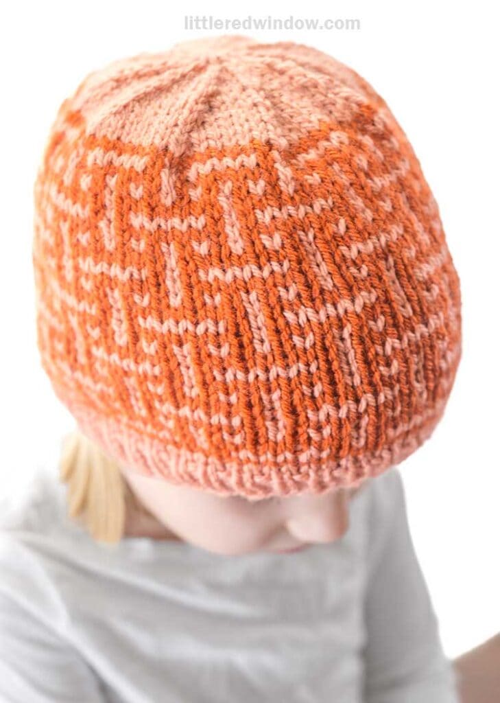 A child wearing an interwoven hat showcases a geometric pattern in shades of orange and peach. With eyes cast downward, the child is dressed in a light-colored top, adding an air of gentle charm to this serene moment.