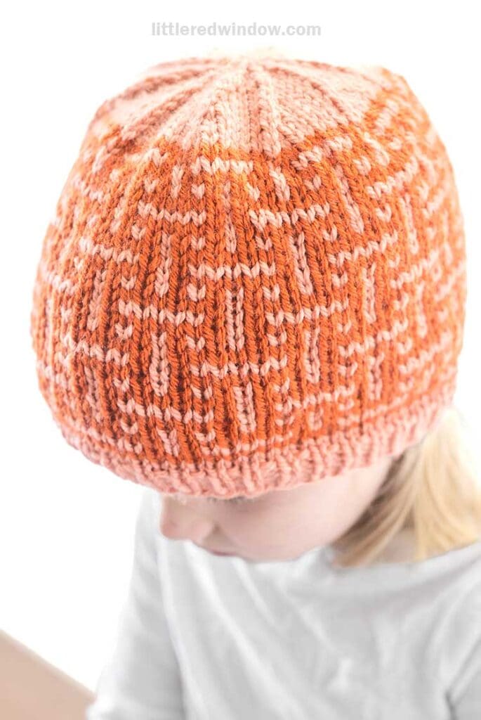 A child wearing an interwoven, hand-knitted winter hat in vibrant orange and pink with a geometric pattern. The child's light hair cascades softly over a white shirt as they look down, set against a plain white background.