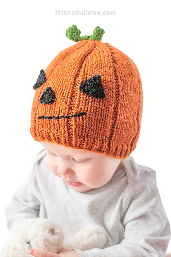 A baby wearing a pumpkin hat, featuring a green stem and black triangle eyes and mouth, sits looking down while holding a stuffed toy. The baby is dressed in a light gray long sleeve shirt.