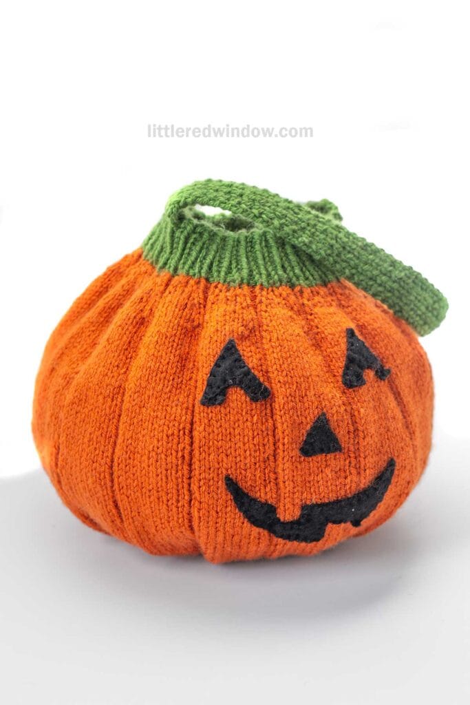 A knitted Jack-o'-Lantern Trick-or-Treat Bag shaped like a pumpkin, featuring a green handle and a black Jack-o’-lantern face, sits on a white background.