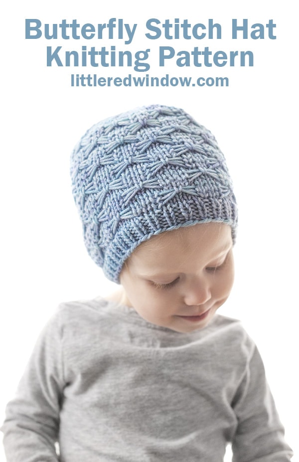 smiling little girl looking down and wearing a light blue knit hat with butterfly stitches all over it