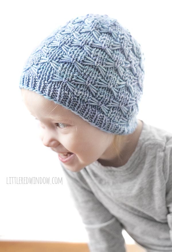 girl laughing and leaning over to the left whitle wearing gray shirt and light blue knit hat with butterfly stitches on it