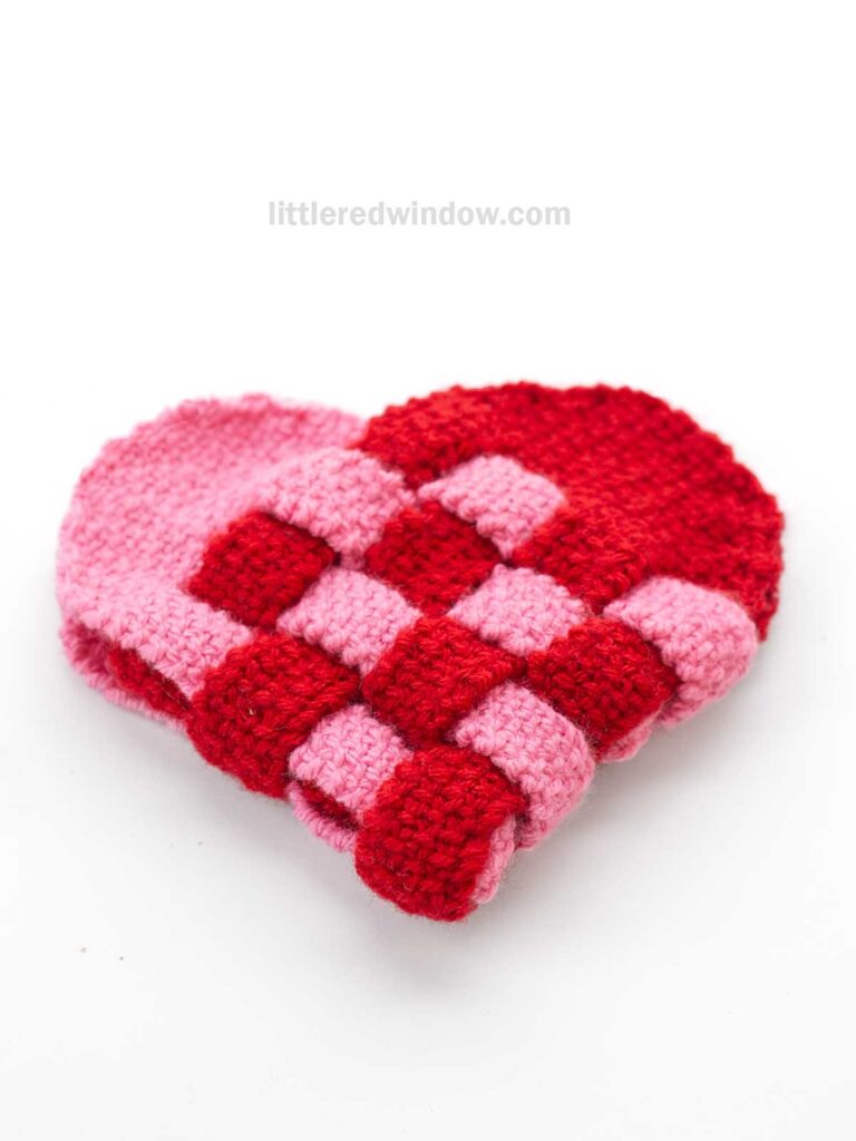 A knit woven heart shape made of interlaced red and pink yarn, creating a checkered pattern. The texture is soft and knit in linen stitch, displayed against a plain white background.