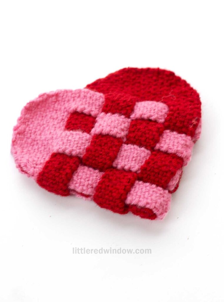 A knitted heart (julehjerte) featuring a red and pink checkered pattern, resting on a white background.