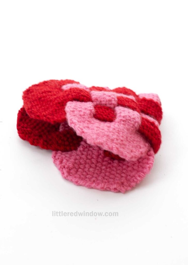 A knitted heart pattern composed of interwoven pink and red segments, that opens up like a pocket, displayed on a white background.