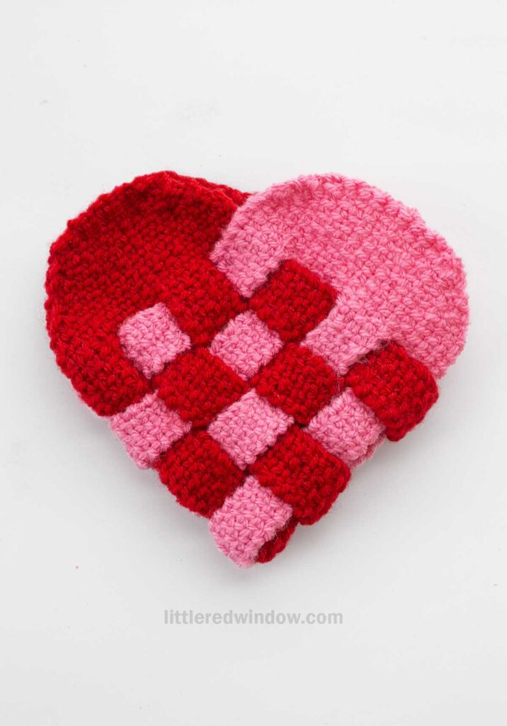 A heart-shaped woven design made from red and pink knit yarn, featuring a checkerboard pattern. The design appears textured and is set against a plain white background.