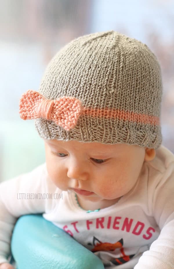 chubby baby looking down and wearing knit gray hat with pink stripe and pink bow on the front