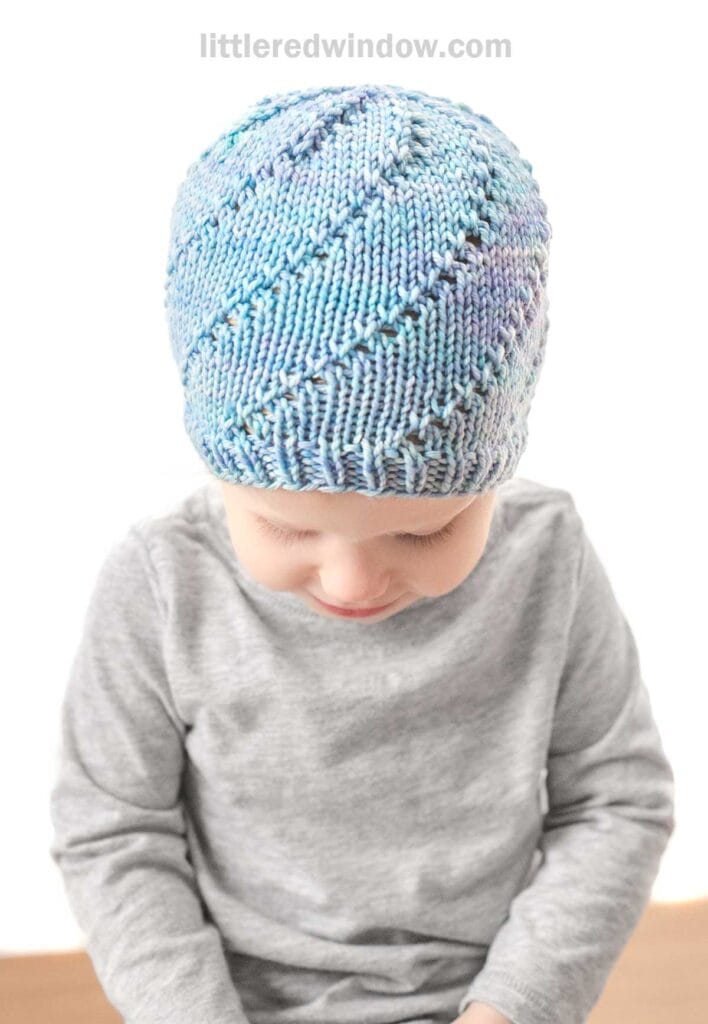 A small child wearing a light gray long-sleeve shirt looks down, showcasing a light blue, hand-knit Lace Twist Hat with a textured swirl pattern. The background is plain, putting full focus on the child and the hat. The words