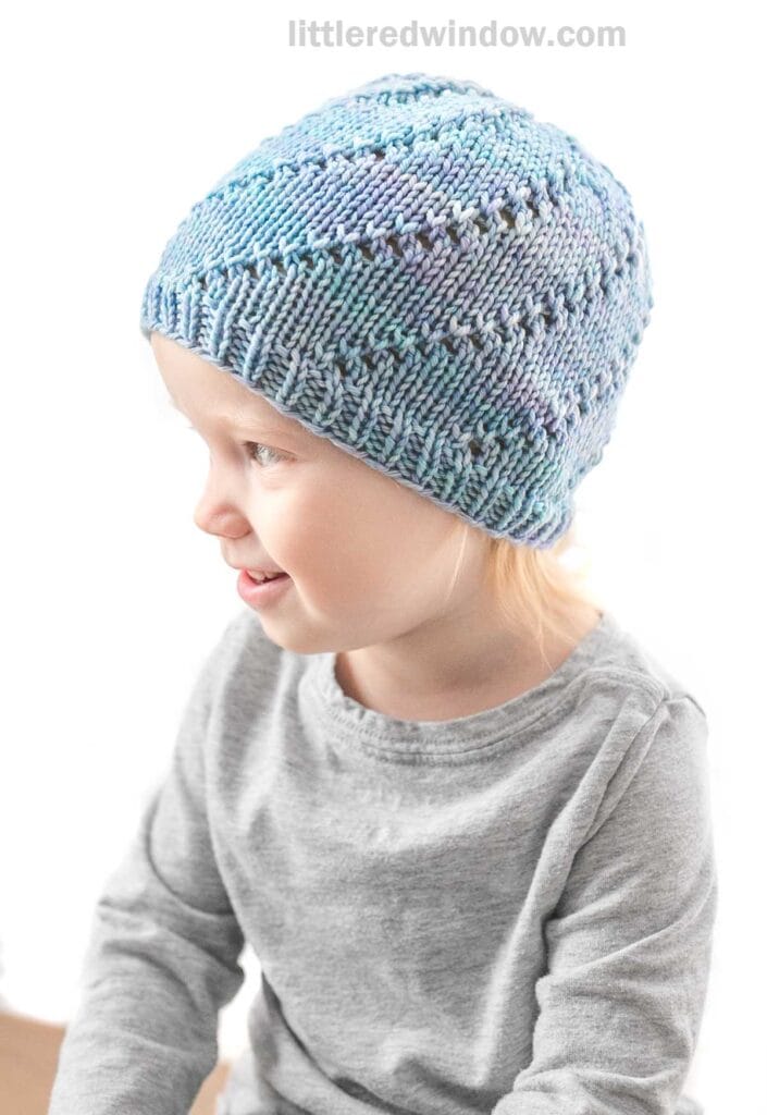 A young child wearing a light blue Knitted Lace Twist Hat with a textured pattern looks to the right, smiling. The child is dressed in a light gray long-sleeve shirt, with light-colored hair peeking out from under the hat. The white background is artfully blurred.
