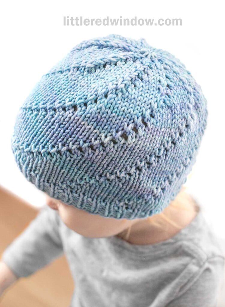 A young child wearing a light blue Lace Twist Hat with a textured pattern sits facing slightly downward. The child is dressed in a gray shirt, and the background is softly blurred. The text