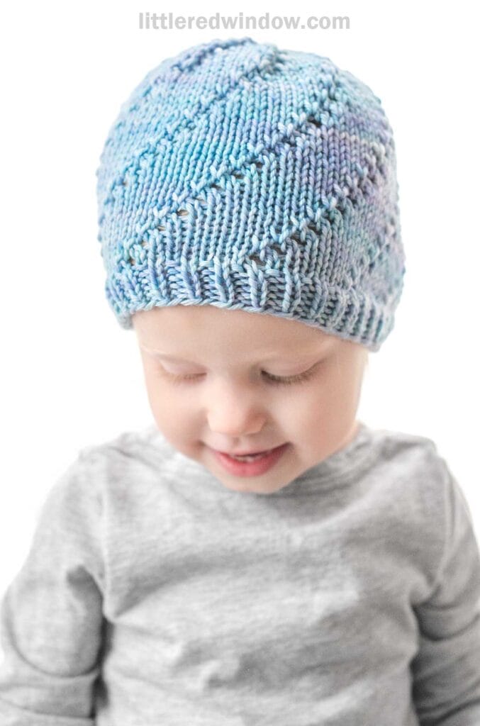 A young child wearing a knitted light blue and purple Lace Twist Hat looks down with a gentle smile. The child is dressed in a gray long-sleeve shirt. The background is plain white, and the top of the image includes the text
