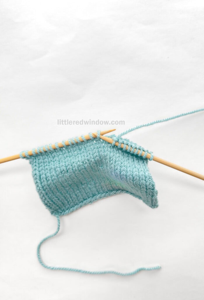 two wood knitting needles and a blue knit stockinette swatch in front of a white background
