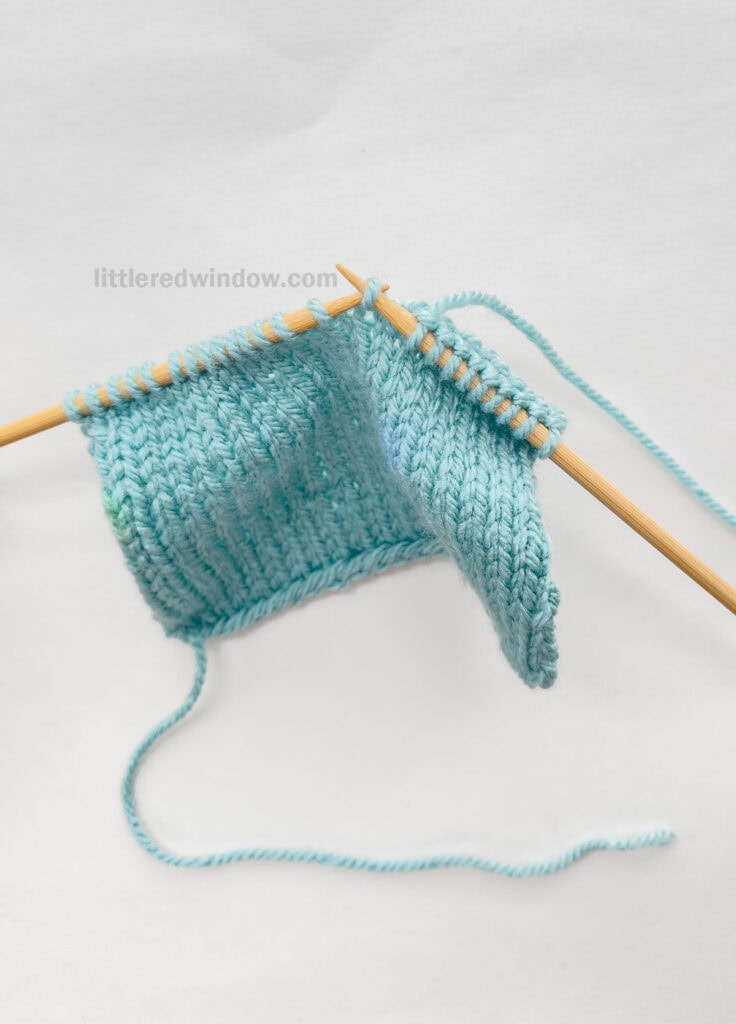 two wood knitting needles and a blue knit stockinette swatch in front of a white background