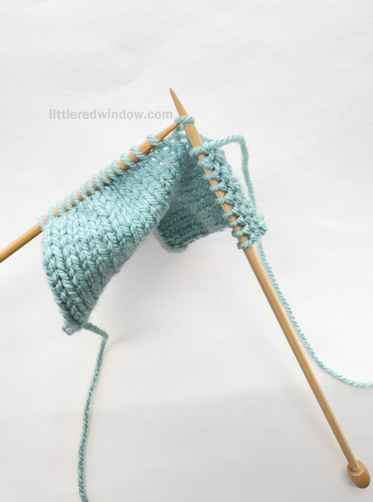 two wood knitting needles and a blue knit stockinette swatch in front of a white background