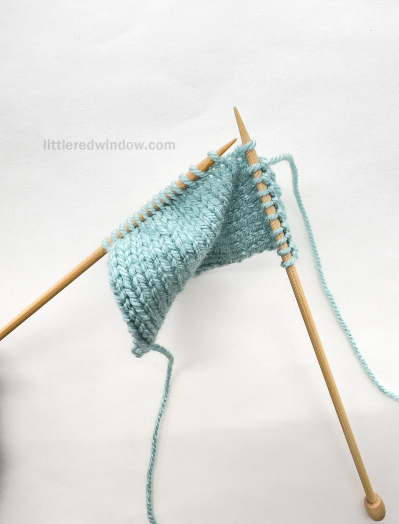 two wood knitting needles and a blue knit stockinette swatch in front of a white background