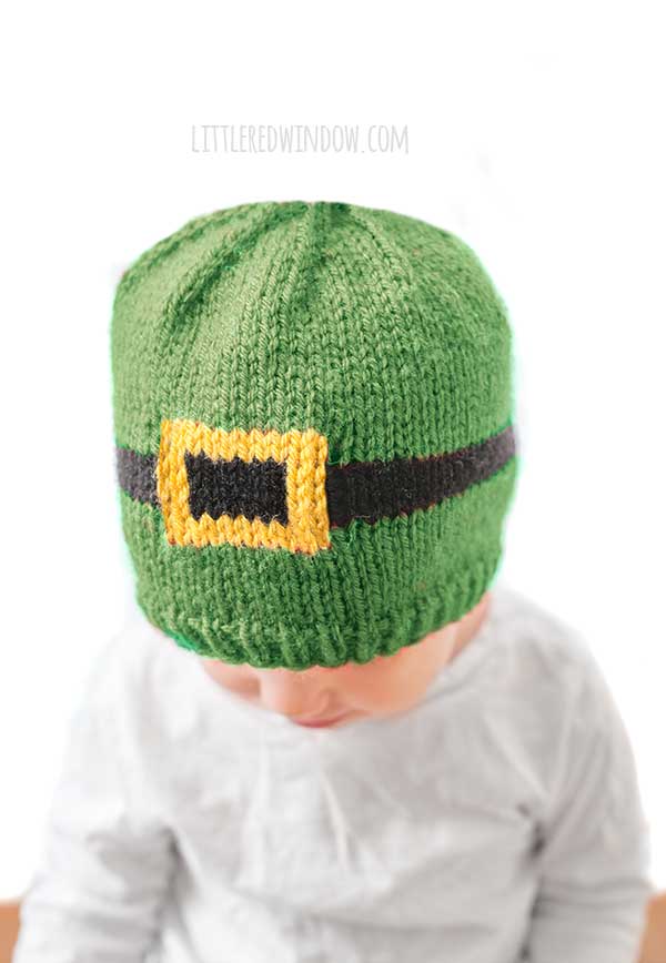 view from slightly above of baby in white shirt wearing a green knit hat with a black and gold leprechaun belt around the middle in front of a white background looking down and to the left